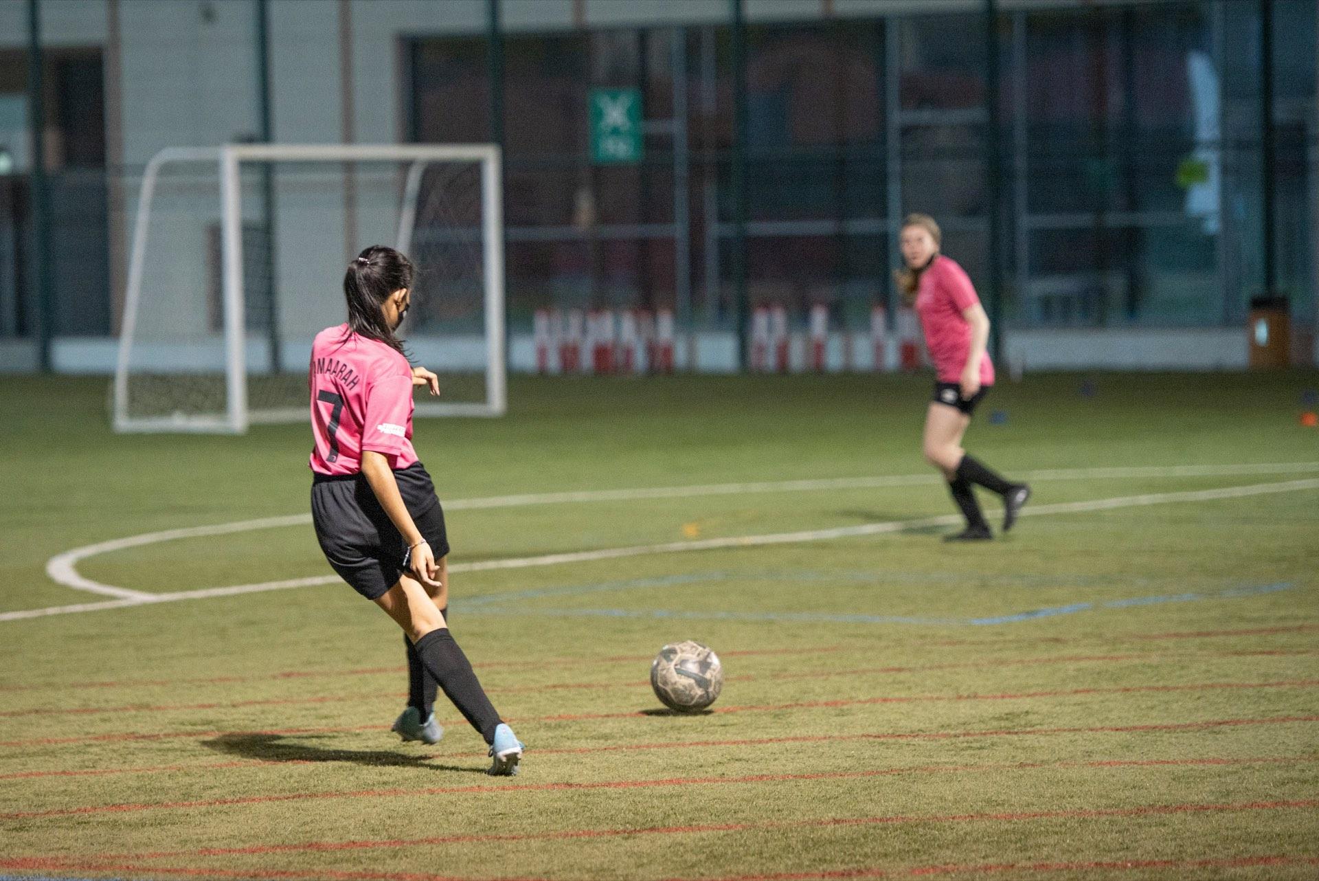 Jugadora de camiseta rosada a punto de patear el balón en una cancha sintética de noche, con una compañera atrás